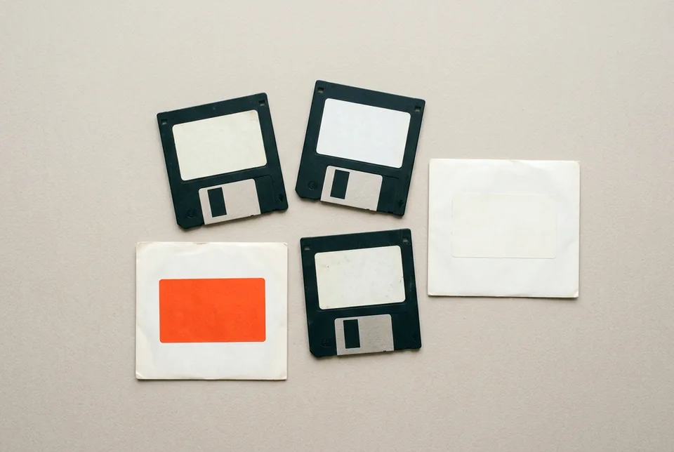 Three floppy disks with blank labels and two envelopes, one with an orange-red label, on grey paper